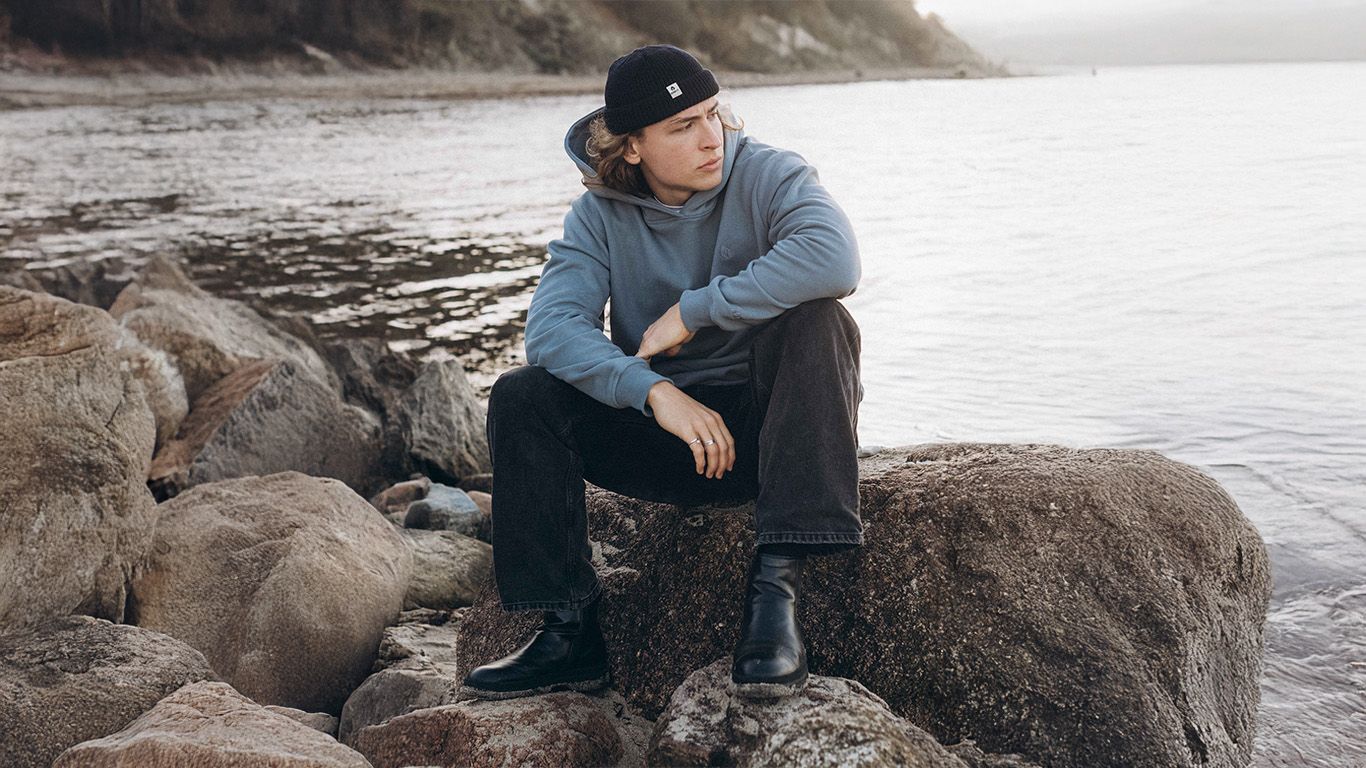 Junger Mann sitzt am Ufer auf einem Stein und guckt in die Ferne des Meeres. Er trägt ein cooles Outfit von Noorlys in marinen Tönen. 