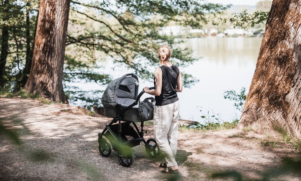Frau spaziert mit Kinderwagen und Baby am See in der Natur.