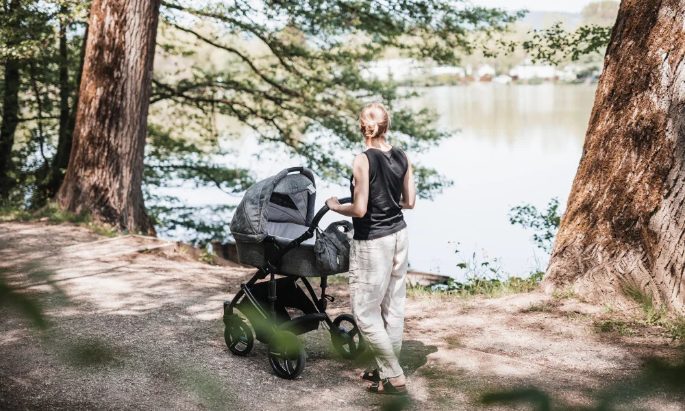 Frau spaziert mit Kinderwagen und Baby am See in der Natur.