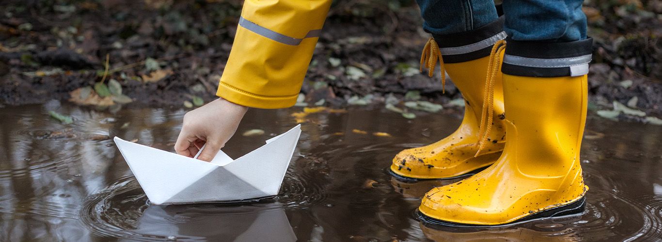 Ein Kind steht in einer sich reflektierenden braunen Regenpfütze mit leuchtenden gelben Gummistiefeln und setzt ein Papierboot aufs Wasser.