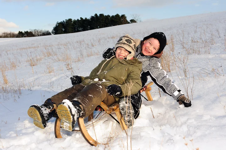 Zwei Kinder fahren Schlitten und lachen. Sie haben dicke Winterkleidung an, angepasst an die natürlichen Wetterbedingungen ist ihr Tag gesichert.