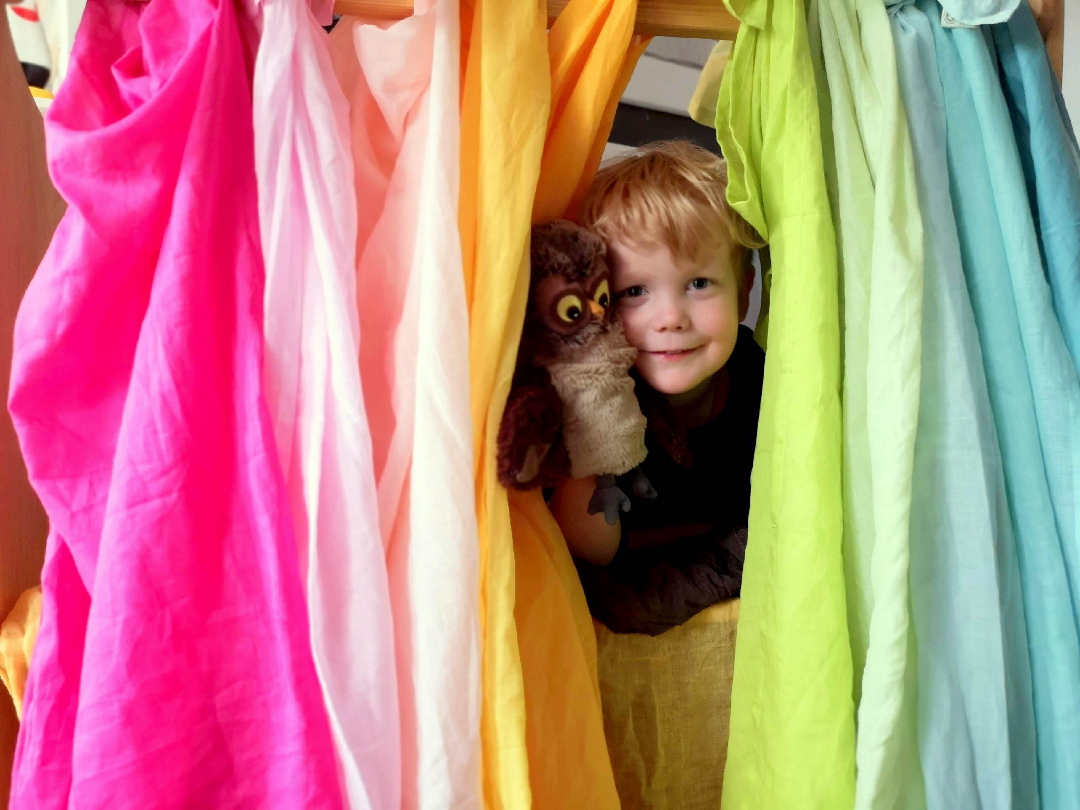 Kleines Kind versteck sich hinter Spieltüchern, die den Eingang für die Höhle darstellen.