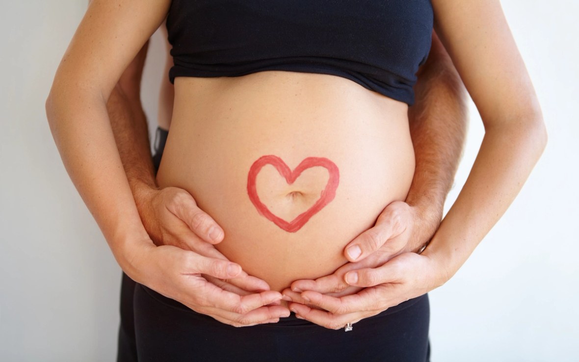 Schwangere Frau wird von hinten umarmt und trägt ein Herz auf ihrem Bauch