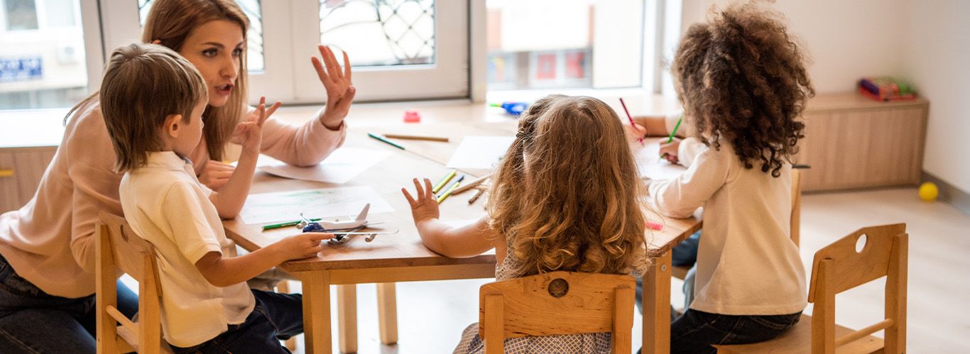 Erzieherin bringt Kindern rechnen bei. Alle sitzen zusammen am Tisch mit Stiften und Zetteln.
